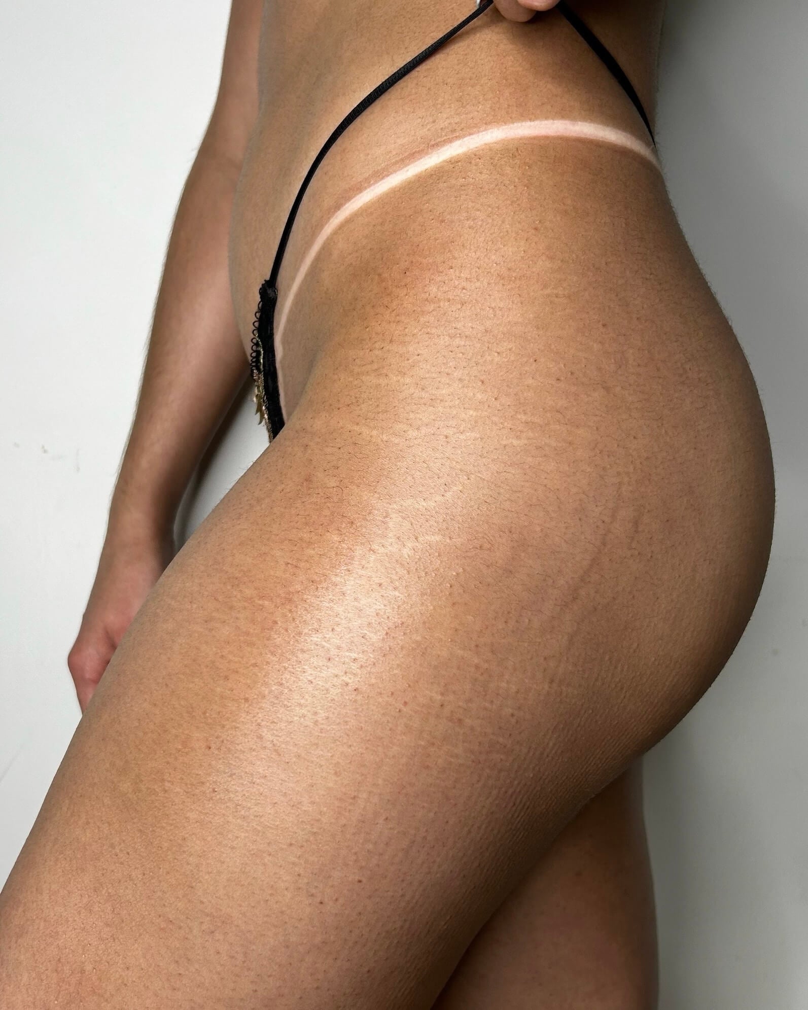 Partial view of a person wearing a black thong against a white background showcasing fake tan tan-lines